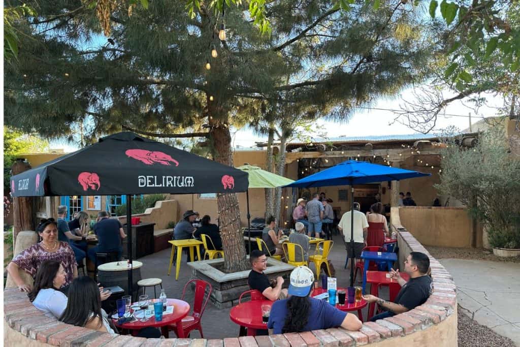People sit at colorful tables under umbrellas in an outdoor patio area surrounded by trees and string lights, enjoying one of the vibrant Las Cruces events, with more people gathered near a lively bar in the background.
