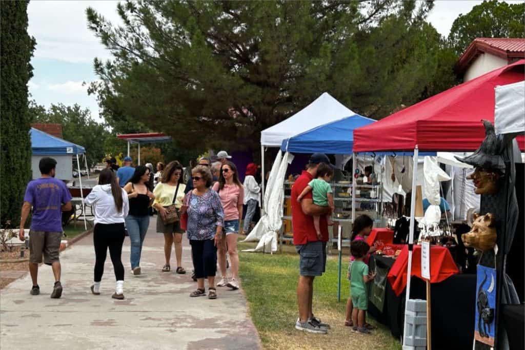 People walk and browse vendor booths at an outdoor market with white, blue, and red tents set up along a sidewalk and grassy area—experience Local Events Las Cruces has to offer.