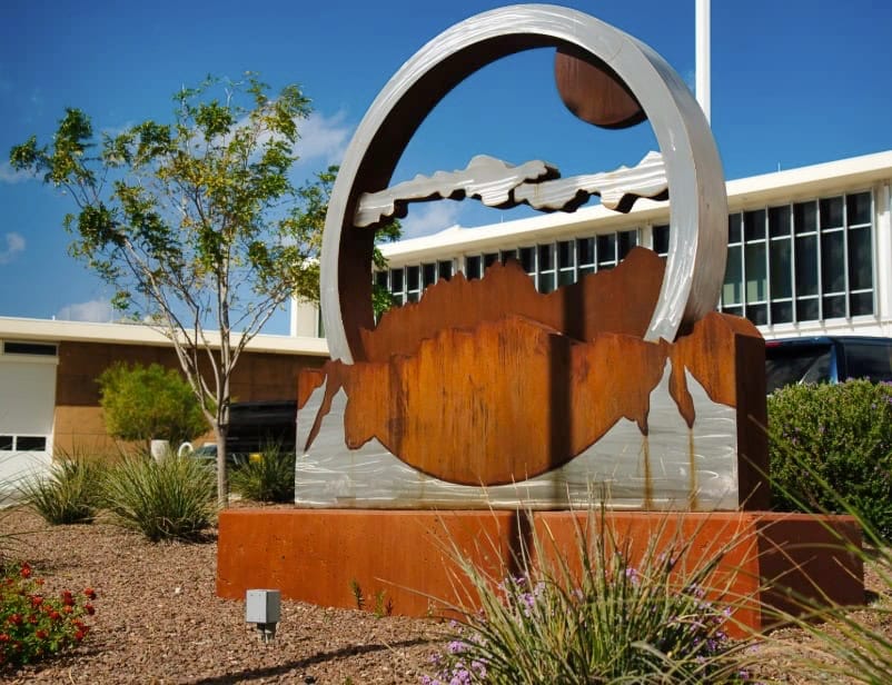 Metal sculpture featuring cloud and mountain cutouts displayed near a modern building in Las Cruces, NM public art area.