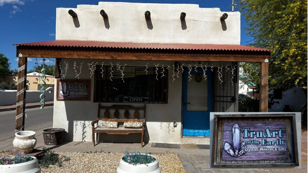 A small Mesilla crystal shop with a wooden bench, potted plants, and a sign reading “TruArt of the Earth Crystals, Minerals & Gifts” under a covered porch; string lights glow above displays of healing stones.