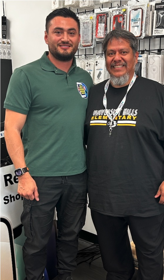 Two men stand smiling in a Las Cruces, NM cell phone repair shop, with phone accessories displayed behind them.