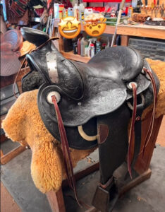 High Noon Leather black tooled western saddle on a stand in Las Cruces workshop, two emoji stickers above.
