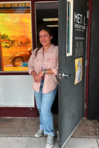 Woman smiling in doorway of Meta'l Jewelry Studio, Las Cruces, wearing jeans and a pink shirt, representing creativity.